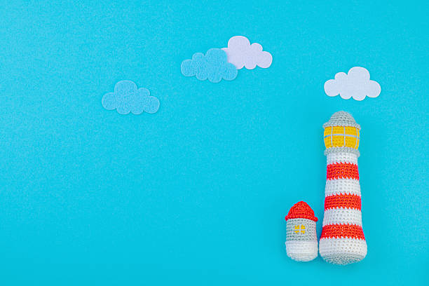 Handmade crochet lighthouse on blue background with felt clouds. Minimalist nautical-themed flat lay with soft textures and vibrant colors, top view