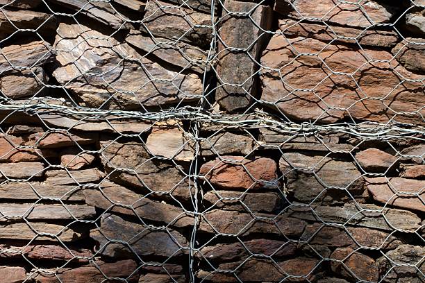A wire fence is wrapped around a stone wall. The fence is made of metal and is covered in barbed wire. The fence is attached to the wall and is not visible from the outside