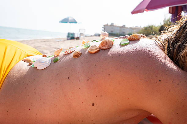 Woman is lying on a sandy beach with various seashells and pieces of sea glass resting on her back, enjoying the summer sun and the relaxing atmosphere