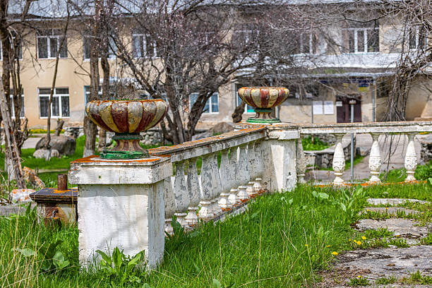 Two ornate Decaying soviet era stone planters display weathered charm in an overgrown garden. Dried plant material adds to the tranquil, aged atmosphere near a building during the daytime.