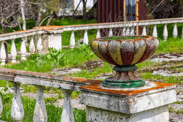 Decaying soviet era ornate planter contains dried plants and moss, showing its age with faded multicolored paint. Stone balustrade sections create a serene, weathered atmosphere in a neglected garden.