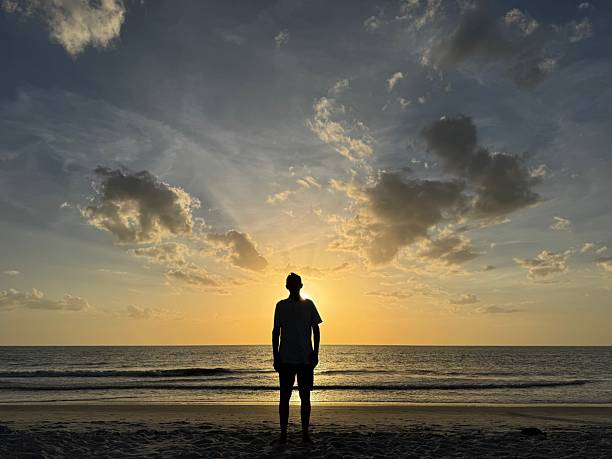 istock Man in silhouette stand boldly on beach in front of setting sun with sun rays seeming to radiate upward from him. 2221910988
