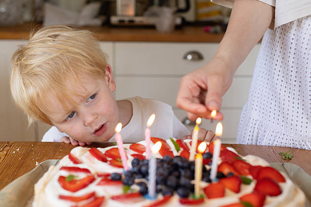 A young blond child looks at lit birthday candles on a pavlova cake with curiosity. A warm and candid family moment in a cozy home.