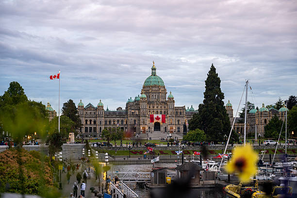 The image displays the British Columbia Parliament Buildings, a prominent landmark situated along the Inner Harbour in Victoria, British Columbia, Canada. A Canadian flag flies from a flagpole on the left side of the building, and a large Canadian flag is draped over the entrance archway. The foreground shows a bustling waterfront area with boats docked in the harbor, and people are visible walking along the waterfront promenade. A bright yellow flower is blurred in the immediate foreground, adding a touch of color to the scene. The sky above is cloudy, suggesting an overcast day or evening.