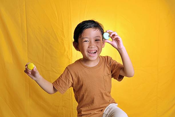 A cheerful boy smiles brightly while showing colorful macarons in both hands, standing against a vivid yellow backdrop in a studio setting.