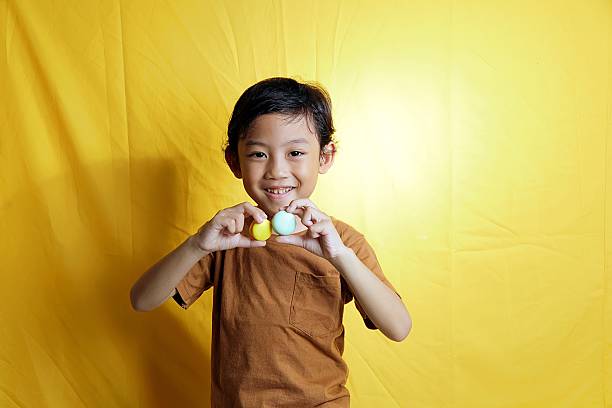 Happy Asian boy holding bright yellow and blue macarons while smiling in front of a vibrant yellow backdrop, expressing joy and childhood fun.