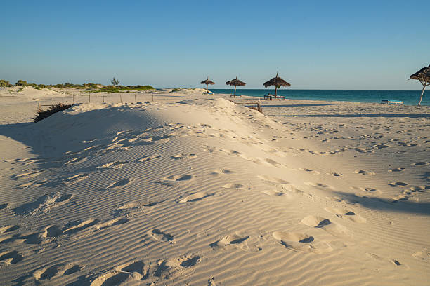 Small dune on White sand beach with people relaxing under umbarella shadows and blues water and sky in background. It is north of Toliara, at Ambatomilo lagoon in South West Madagascar.