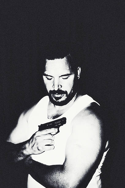 High-contrast black and white portrait of a man looking down while holding a handgun against his chest. The dramatic lighting and heavy shadows evoke a moody, introspective atmosphere. The subject’s expression suggests inner conflict or contemplation. Visual style resembles gritty film noir from the 1970s–1990s.