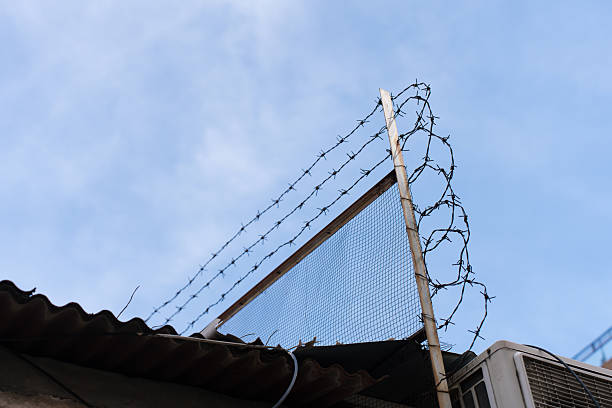istock Rusty iron mesh fence with barbed wire on top of a building under blue sky. 2222016475