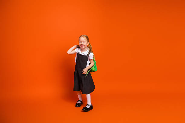 istock Happy young girl wearing school uniform posing with her backpack on an orange background, representing education 2222018410