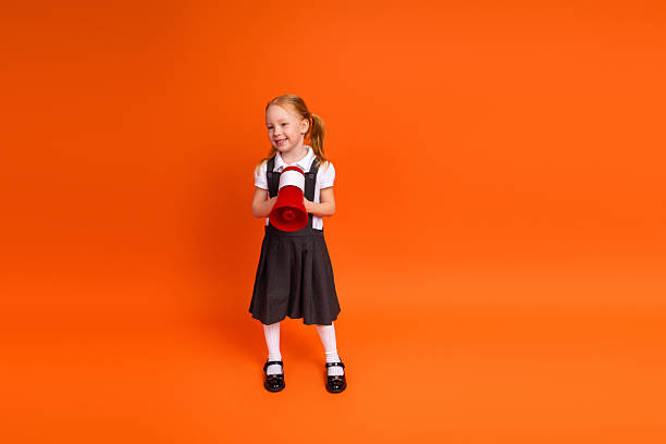 istock Cheerful schoolgirl in uniform holding red megaphone while posing on vibrant orange background 2222018461