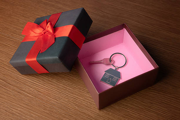 An open gift box with a red ribbon reveals a house key and a house-shaped keychain inside. New home, homeownership, celebration, gifting concept.