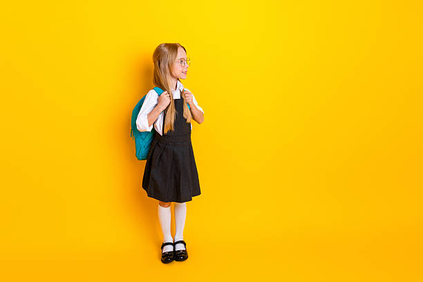 istock Charming elementary schoolgirl in uniform with backpack stands against bright yellow background, ready for the first day of school 2222026540