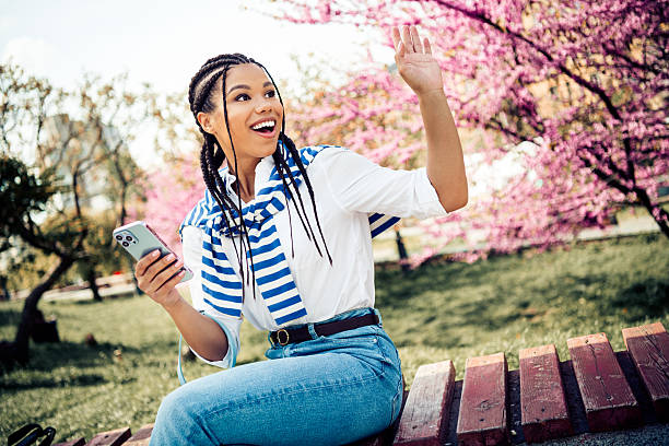 istock Charming woman enjoying a spring day outdoors in a blooming park, smiling and waving with her phone in hand 2222039921