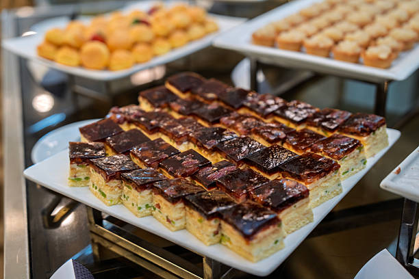 Close-up of layered egg squares topped with sausage, neatly arranged on a buffet platter with other assorted canapes in the background.