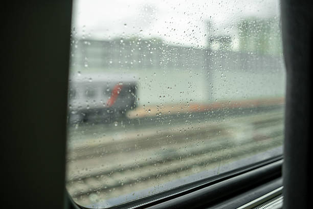 Raindrops cling to a train window while the vehicle passes through a city area, showcasing the blurred scenery outside on a grey day.