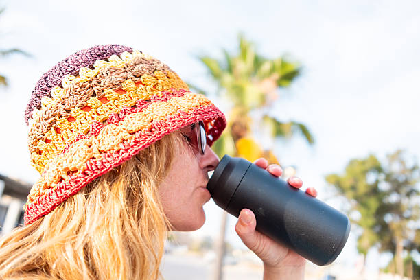 Blonde tourist with colorful crochet hat and sunglasses drinking a refreshing drink from a black thermos on a sunny summer day, with palm trees and blue sky in the background