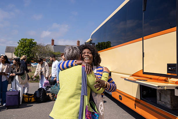 Two senior women friends reuniting in front of a coach, hugging each other. They are visiting the North East of England, excited for their trip.  Other passengers are standing around them. They laugh while they hold each other.Videos are available similar to this scenario.