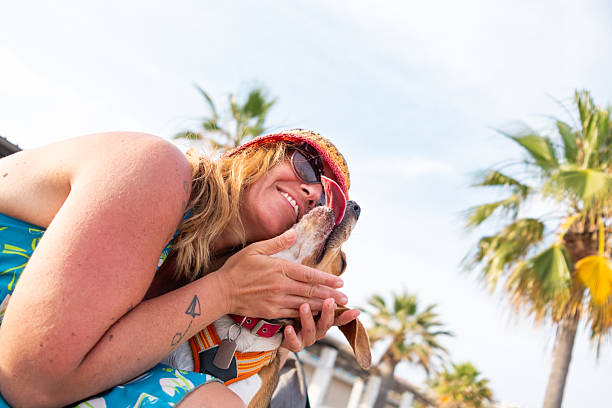 Smiling blonde woman wearing sunglasses and summer hat embracing her beagle dog on a sunny beach with palm trees in the background, enjoying summer vacation together