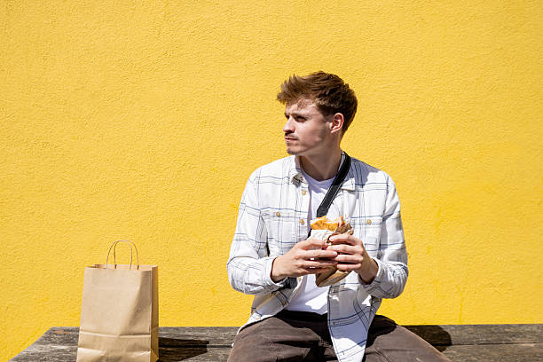 Medium shot of a young adult male sitting in front of a bright yellow wall in Newcastle-upon-Tyne, North East England. He is looking off-frame while enjoying a sandwich on his lunch break. There is a camera around his neck and brown paper bag on the bench beside him.\n\nVideos similar to this scenario available.