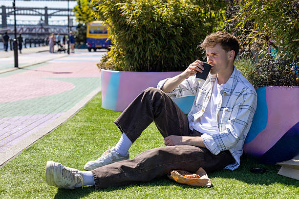 Medium shot of a young adult male drinking a coffee while sitting in a city centre pedestrian area in Newcastle-upon-Tyne, North East England. He is wearing casual clothing and the sun is shining. There is a pastry on a paper bag beside him.\n\nVideos similar to this scenario available.
