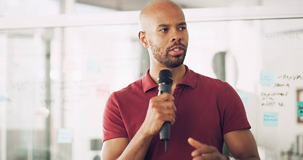istock Presentation, speaker and man with mic, talking and information for seminar, training and creative. Business, employee and black person with speech for workshop, conversation and public speaking 2222069281