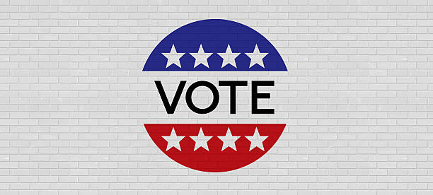 A classic American political campaign badge is featured prominently against a white brick wall. The red, white, and blue design, complete with stars and the bold word \