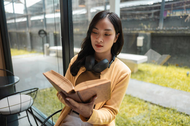 Asian student enjoying a captivating book while relaxing in a cozy cafe, surrounded by the warm glow of natural light and urban ambiance