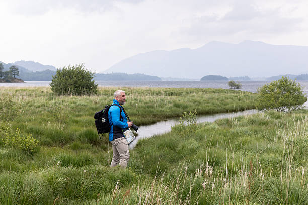 A wide shot of a male photographer, dressed in outdoor gear, exploring a lush grassy wetland with a camera in hand and a backpack. The scene features tranquil water, dense greenery, and misty mountains in the background under an overcast sky. He is also carrying a map to navigate around the area.\n\nVideos are available similar to this scenario.