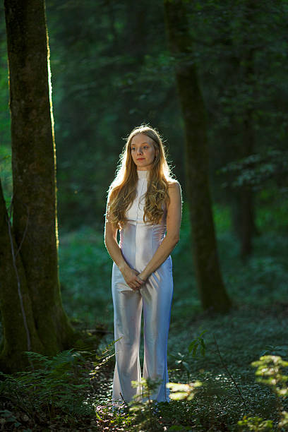A serene and elegant young woman in a white jumpsuit standing quietly in a sun-dappled forest. With long flowing hair and a composed expression, she is surrounded by tall trees and green foliage. A calm and introspective moment captured in natural light, symbolizing peace, stillness, and connection with nature.
