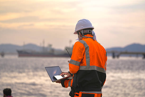 istock Two engineers wearing safety uniforms and hard hats communicate using a radio while reviewing data on a laptop by the sea during sunset. Maritime industrial setting. 2222107659
