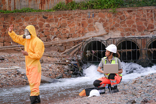 Environmental field team in protective gear analyzes water samples on a polluted beach. One holds a laptop while the other handles vials for chemical testing of coastal pollution.