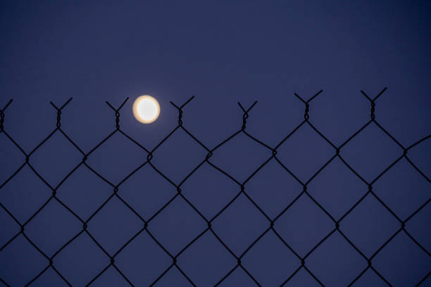 A fence with a moon in the sky. The moon is the only light in the image. The fence is black and white