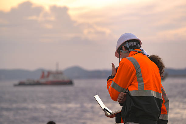 Two engineers wearing safety uniforms and hard hats communicate using a radio while reviewing data on a laptop by the sea during sunset. Maritime industrial setting.