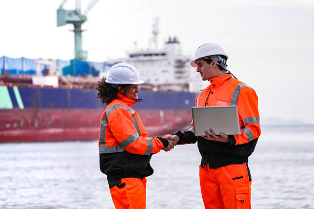 istock Two port engineers in high-visibility orange uniforms and hard hats communicate while inspecting dockside logistics using a laptop and walkie-talkie, symbolizing teamwork and safety. 2222110333