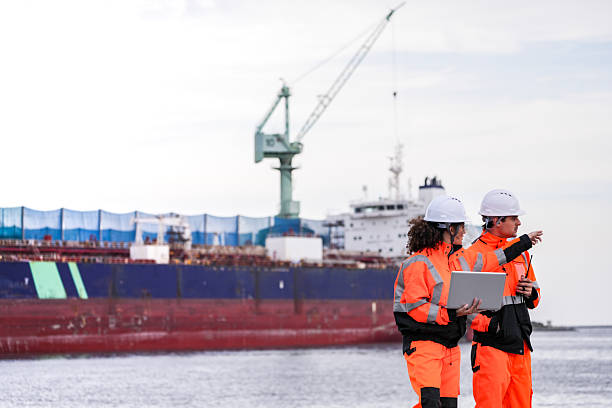 istock Two port engineers in high-visibility orange uniforms and hard hats communicate while inspecting dockside logistics using a laptop and walkie-talkie, symbolizing teamwork and safety. 2222110403