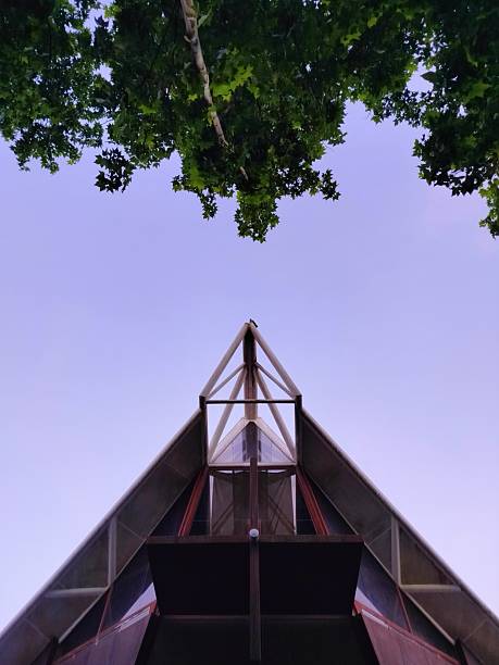 An upward view of a modern triangular church facade framed by leafy branches and a lavender sky at dusk. The structure features angular lines, large glass panels, and a prominent metal framework, creating a striking sense of symmetry and architectural clarity. The interplay between nature and built form evokes a contemplative atmosphere, blending spiritual design with geometric precision.