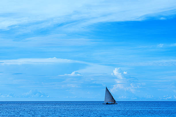 A small sailboat navigates peaceful blue waters beneath an expansive sky filled with white clouds, showcasing the serene beauty and freedom of the open sea.
