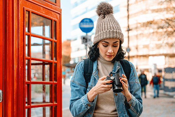 istock Young woman examines camera settings while standing next to red telephone booth in urban setting on a sunny day 2222120696