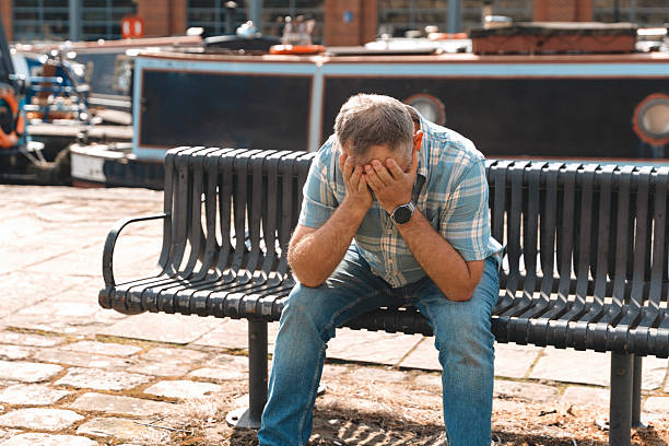 A man dressed in casual attire sits on a bench beside the water, covering his face with his hands. It is a bright afternoon, and he appears deep in thought.