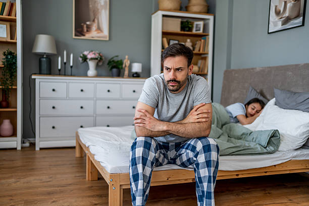 istock Frustrated man sitting on bed while wife sleeps peacefully 2222123362
