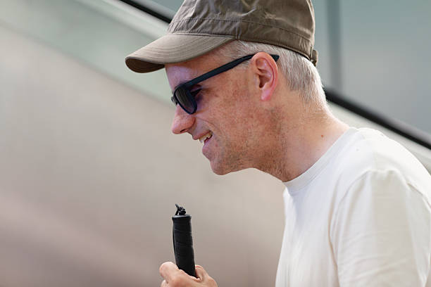 Portrait of mid adult smiling blind man, wearing white shirt, green cap and sunglasses, holding blind persons cane, profile view