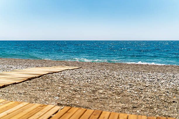 A low-angle view of a simple wooden walkway cutting through a pebble-covered beach, leading to the calm, glittering sea. The sky is perfectly clear with no clouds, adding to the serene and untouched feel of the landscape. Ideal representation of peaceful beach access.