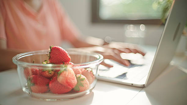 Asian young woman enjoying strawberries from a reusable food container while using laptop.