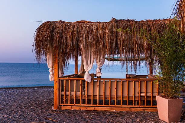 Cozy wooden cabana on a pebble beach, featuring a roof made of dried palm leaves and flowing white curtains tied neatly at the sides. The cabana faces the calm sea under a soft, dusky sky, evoking relaxation and exclusivity in a beach resort setting.