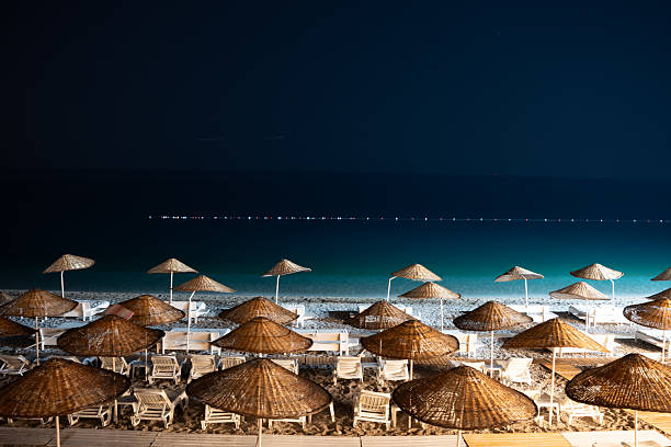 A night capture of a serene beach, showcasing organized rows of empty loungers and thatched umbrellas. The gentle glow from lights highlights the textures of sand and water, while the dark sea blends into the infinite night sky in the distance.
