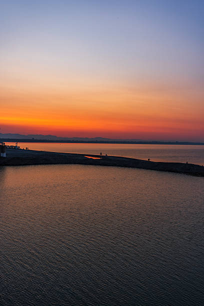 Vertical shot of a golden-orange sunset reflecting on a small lagoon next to the sea. People are seen in silhouette walking along a rocky pier, creating depth and scale. Distant mountains and coastal lights add atmosphere to this calm yet vivid composition.