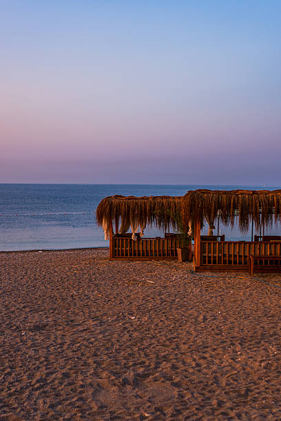 A more distant view of cozy wooden cabanas aligned neatly along the shoreline. The soft pastel colors in the sky reflect a tranquil, post-sunset atmosphere. Ideal for themes of resort lifestyle, relaxation, and seaside architecture.