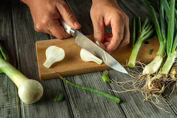 istock Hands skillfully cut fresh green onions on a rustic wooden board. The vibrant vegetables include roots and crisp green tops, ideal for enhancing dishes. Culinary activity unfolds in a cozy kitchen 2222138763