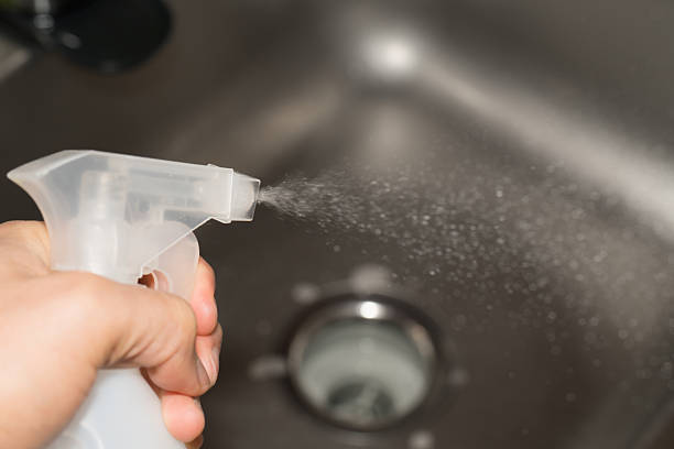 A hand presses a cleaning spray bottle, releasing a fine mist of cleaning solution into a stainless steel kitchen sink. The image captures the dynamic motion of droplets in mid-air, representing cleanliness, hygiene, disinfection, and household chores. Ideal for use in concepts of domestic care, sanitation, and lifestyle routines